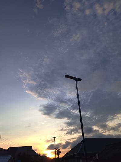 This image captures a twilight scene in Rumuokwuta, Nigeria. The sky is a gradient of pale blue and soft grays, with scattered clouds, some appearing wispy and others more dense. A sliver of the moon is visible in the upper portion of the sky. Dominating the mid-ground is a tall, dark lamppost with a rectangular solar panel light fixture at its apex, casting a warm, orange glow. Below the lamppost, silhouetted against the horizon, are the dark roofs of several buildings, suggesting a residential or urban area. The setting sun is visible on the left side of the frame, a bright orb of yellow and orange peeking through the clouds, casting a warm light on the lower sky. Other smaller light poles are also present in the distance. The overall impression is one of evening settling over the city. There are no people or animals visible in the frame.