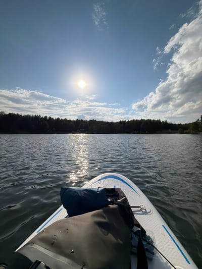 The image shows a view from a paddleboard on a body of water, likely a lake or river. The paddleboard is white with blue accents and has a dark-colored bag attached to it. The water has visible ripples and reflects sunlight, creating shimmering highlights. In the background, there is a treeline indicating the shoreline. The sky is blue with scattered white clouds and the sun is visible. While the prompt mentions Selychka, Russia, there is no information in the image that confirms that this is the location.