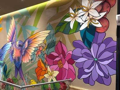 The image displays a detailed, stylized mural on an interior wall in Santa Tecla, El Salvador. The mural features tropical flora and fauna against a light beige background.
A prominent, multicolored bird, characterized by iridescent blue, purple, orange, and pink hues across its body and large, outstretched wings, occupies the central left portion of the mural, depicted in mid-flight and facing right. A section of a smaller, light blue bird is visible at the lower-left edge.
The upper-right section of the mural presents white coffee blossoms with yellow pistils, alongside clusters of red coffee cherries and dark green leaves. Below these, a large, multi-petaled purple flower is depicted, occupying the lower-right quadrant. To its left, a similarly large, multi-petaled pink/magenta flower is present. Smaller orange and white flowers are visible towards the lower-center. Various light green and teal foliage elements are integrated throughout the mural's background.
A linear light fixture is installed along the top edge of the wall, providing indirect illumination. In the lower-left foreground, a silver-colored metal railing, consistent with a staircase or barrier, partially obscures the mural. A white rectangular wall vent or fixture is visible on the far right edge of the wall, above the purple flower. No individuals or dynamic actions are present in the scene.