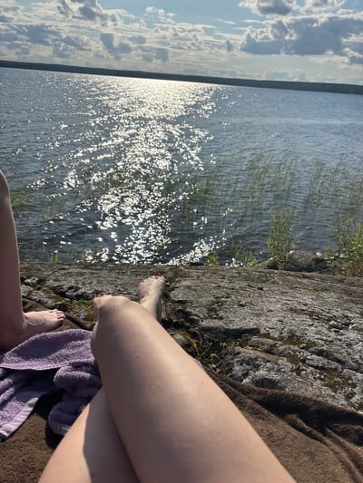 The image shows a close-up of two pairs of bare legs and feet resting on a rocky shore next to a body of water. A purple towel is visible near the left leg. The water is calm and reflects the sunlight, with reeds growing along the edge of the shore. In the distance, a tree-lined horizon is visible under a cloudy sky. The scene suggests a summer day spent outdoors by the water. The specific location is the shore of Lake Ladoga in Shuiskaya Chupa, Russia.