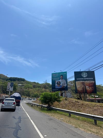 The image shows a road scene in Pozos, Costa Rica, specifically at the coordinates 9.948 latitude and -84.178 longitude. The road is a two-lane highway with a white dividing line. The asphalt surface is dry and appears to be in good condition. A silver sedan is traveling in the lane closest to the viewer, while several other cars are visible in the distance, including a pickup truck with a blue tarp over its bed. The road curves slightly to the right, and on the right side of the road, there is a grassy embankment topped by a metal guardrail. There are two billboard signs visible on the embankment. The sign on the left advertises DHL, featuring a white male with dark hair and a blue shirt. The sign on the right advertises Shaken Espresso, featuring a product of two glass cups filled with brown liquid. Green trees and vegetation are visible on the hillside in the background. The sky is clear and blue with some wispy white clouds. The image was likely taken during the daytime, as the lighting is bright and there are no shadows.