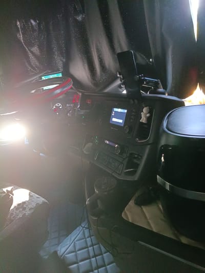 The image shows the interior of a truck cabin, with a focus on the dashboard and driver's seat area. A bright light source, possibly sunlight, illuminates the left side of the image, casting strong glare on the steering wheel and dashboard. The dashboard features various instruments and screens, including a navigation display and a radio. A phone is mounted on a holder on the right side of the dashboard. A small white figurine is visible on the dashboard, near an air vent. The driver's seat on the left is partially visible, with a patterned fabric covering. The floor is covered with quilted mats. The overall environment appears to be indoors, within a vehicle. The location is identified as Uttrichshausen, Germany.