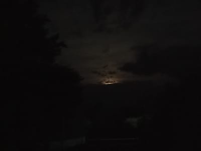 The image depicts a nighttime scene in Mykhnivtsi, Ukraine, with a partially obscured moon visible through clouds. The foreground is dominated by darkness, with a hint of what appears to be a building or structure in the lower left quadrant. Silhouetted trees line the left side of the frame, contributing to the overall dark and somewhat ominous atmosphere. The moon itself is a bright, diffused light source peeking through the cloud cover, suggesting it is either a full or gibbous moon. The environment appears to be rural or suburban, given the presence of trees and the apparent lack of significant urban illumination.