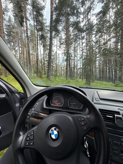 The image depicts the interior of a BMW vehicle, viewed from the driver's perspective. The steering wheel, dashboard, and a portion of the car's interior are visible. Through the windshield, a dense forest of tall pine trees and green undergrowth is seen, suggesting a rural or natural setting, likely on the outskirts of Yundola, Bulgaria. The weather appears overcast. There are no people visible in the image. The car is stationary.
