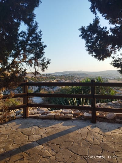 The image shows a scenic overlook in Għargħur, Malta, at sunset. A wooden fence separates a paved stone patio from a valley below. The patio is made of large, irregular flagstones with visible cracks and shadows cast by unseen objects. Behind the fence, the valley is covered in rocky terrain, sparse vegetation, and some low-lying plants, possibly agave or yucca. In the distance, a town or village with buildings is visible nestled among rolling hills. The sky is clear and a pale blue, transitioning to a lighter hue near the horizon. Dark silhouettes of trees frame the top corners of the image. A timestamp at the bottom right corner reads "05/07/2026 19:50". No people are visible in the frame.