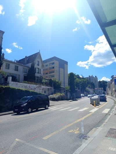 The image captures a street scene in Nancy, France, on a bright, sunny day with scattered clouds. A black van is parked on the left side of the road, facing forward. The street has yellow lane markings and a pedestrian crossing. In the background, there are buildings of various architectural styles, including a multi-story building with a light blue and yellow facade, and a more traditional white building with a pointed roof. Trees line the street and the hills behind the buildings. A bus stop shelter with a glass roof is visible in the upper right corner. The sky is a clear blue, with the sun shining brightly and causing lens flare.