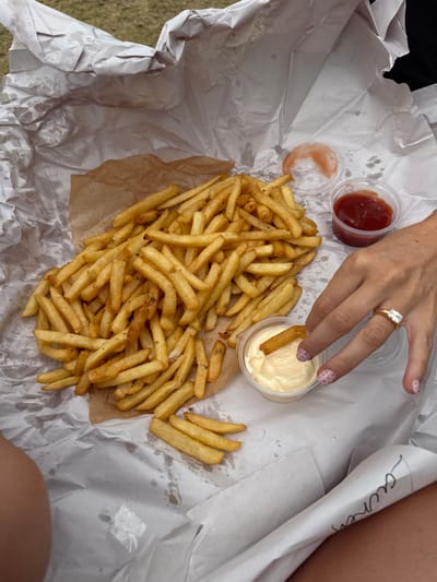 A high-angle shot shows a pile of golden fries on crumpled white paper, accompanied by small plastic containers of ketchup and mayonnaise. A hand with a prominent diamond ring dips a fry into the mayonnaise. Beside the fries, a paper wrapper shows a written name. The overall setting suggests a casual outdoor meal.