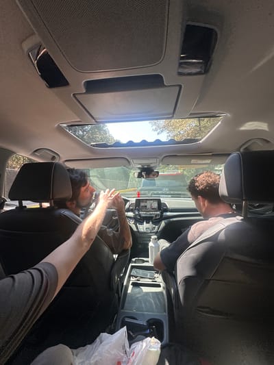 The image, taken inside a vehicle during a bright, sunny day, shows two men in the front seats. The car interior features black seats, a rearview mirror, and a dashboard equipped with an infotainment screen and a small device mounted near the windshield.

The driver, with dark, curly hair and a beard, is seen wearing a grey shirt. His arms are extended towards the windshield, holding a yellow object, possibly a smartphone or another device. His face is partially visible, showing concentration. The passenger, whose back of the head and shoulder are visible, has short, curly brown hair and is wearing a dark, long-sleeved garment.

A white plastic bag is present in the passenger footwell, and a white bottle with "Qualcomm" text is visible near the passenger side. Through the car windows, bright sky and trees are discernible. A reflection in the rearview mirror suggests a "Hyundai" logo on the infotainment screen. The scene depicts the occupants engaged in activities within a parked or stationary car in what appears to be Coajomulco, Mexico, during daylight hours.