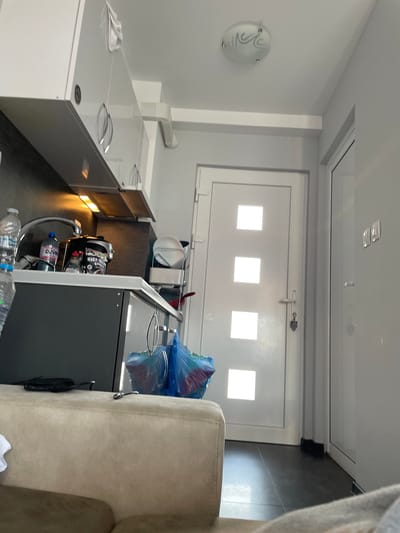 The image depicts an interior view of a small kitchen and a portion of a hallway in an apartment in Chaidefto, Greece. The kitchen features modern, white, high-gloss cabinets with silver handles. Underneath the cabinets, task lighting illuminates the countertop. The countertop itself is a lighter material, and it holds a sink with a chrome faucet, two water bottles (one with a blue label), a coffee maker or similar appliance, and a container labeled "HIGH PROOF GREEN." To the right of the countertop, there is a metal rack with stacked white plates. In the background, a white door with three square glass panes is visible, suggesting it leads to another room or an exit. The door has a silver handle and a key attached to a keyring. To the right of this door is another white door, partially visible, with a silver handle. The floor is dark tile. In the foreground, a beige sofa is partially visible, indicating the space is a living area adjacent to the kitchen. The walls are painted a light gray. A ceiling light fixture with a decorative glass shade is mounted on the ceiling. Two blue plastic bags are on the floor near the kitchen counter.