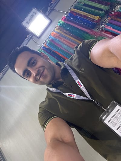 An upward-angled, selfie-style photograph depicts a light-skinned adult male indoors. The subject, positioned centrally, presents a subtle smile and looks directly at the camera. He wears a dark olive green polo shirt with thin light-colored stripes on the sleeves. Around his neck is a grey lanyard featuring repeated "RMC" text in red, green, and blue, connected to a clear plastic identification holder. The badge within the holder displays partially legible text, including sections for "Show Dates & Timings" and dates such as "16.10.19," "17.10.19," and "18.10.19," with corresponding times. The background to the left features a bright, square LED panel light fixture mounted high. To the right, a retail or exhibition display rack is visible, heavily laden with numerous hanging strands of small, sparkling, multi-colored beads or sequins in various hues including blue, green, pink, purple, orange, red, and yellow. A plain, light-colored wall or partition is present in the lower left background. The precise location within Jaipur Municipal Corporation, India, cannot be determined from the visual information provided in the image.