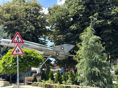In the city of Yoakim Gruevo, Bulgaria, a monument of a fighter jet is displayed outdoors. The jet, painted in a camouflage pattern, is positioned horizontally, suspended by supports. In front of the jet, two triangular road signs are visible. The top sign depicts a pedestrian crossing, while the bottom sign shows two children walking. Both signs are red and white. The area is surrounded by lush green trees, including a large coniferous tree on the right and other deciduous trees behind the jet. A clear blue sky with scattered white clouds forms the background. The foreground features a paved walkway, a decorative metal railing, and some shrubbery. A small, white car is visible parked in the distance.