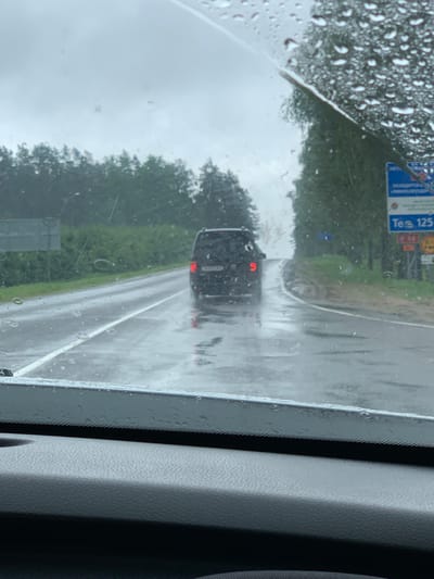 This is a forward-facing, eye-level shot from inside a vehicle, looking through a rain-streaked windshield. The exterior view shows a wet asphalt road with white lane markings, curving to the right and leading uphill. A dark-colored SUV with its brake lights illuminated is driving ahead on the road. Dense green trees line both sides of the road, and the sky is overcast and gray. A blue road sign with white lettering, including the text "Tea 125" and a red circular symbol, is visible on the right side of the road, near the trees. The foreground shows the textured dashboard of the vehicle. The overall scene suggests a rainy day on a rural road in the vicinity of Мядзельскі сельскі Савет, Belarus.