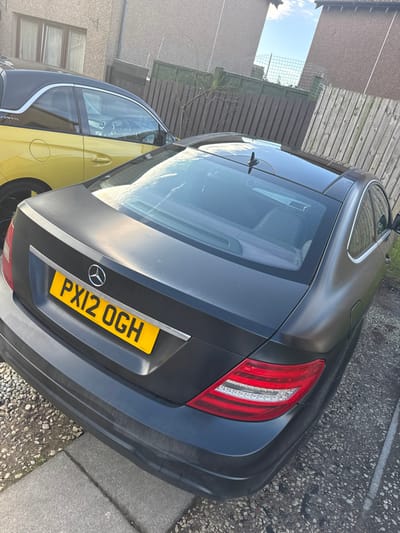 A dark grey matte-finish Mercedes-Benz C-Class Coupe, bearing UK registration plate PX12 OGH, is parked diagonally on a gravel and concrete paving surface. The vehicle's rear is prominent, featuring illuminated taillights with LED strips and a rear bumper exhibiting minor scuffs and dirt accumulation, with integrated parking sensors visible. A black shark fin antenna is centrally mounted on the car's roof. To the left, the rear-side of a yellow two-door vehicle with a black roof, consistent with a Vauxhall Adam model, is partially visible. In the background, a brown vertical wooden slat fence extends across the scene, beyond which bare tree branches are discernible. Further in the distance, parts of two light-colored residential buildings with windows are visible, separated by an exposed section of sky. A metal mesh fence, topped with barbed wire, is present between these buildings. The scene is outdoors, illuminated by natural daylight under a partly cloudy sky. The specific location within Montrose, United Kingdom, cannot be precisely determined from the visual evidence provided.