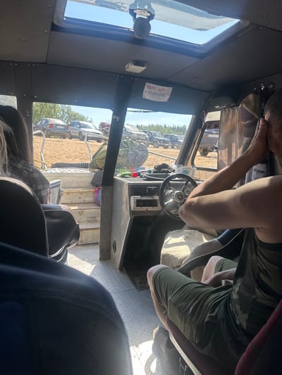 The image, taken from inside a vehicle, likely a boat or an all-terrain vehicle, shows a view through the windshield and side windows. The setting appears to be an outdoor, possibly rural, area with sandy ground and trees visible in the background. Several parked vehicles are present outside, including what appear to be cars and an ATV.
Inside the vehicle, the focus is on the driver's seat and the passenger area. A person is visible in the driver's seat, with their hands on the steering wheel. Another person is seated in the foreground, facing away from the camera. Various objects are scattered within the cabin, including what looks like a steering wheel, dashboard elements, a hanging hat, and a red and blue bag. A piece of paper with Cyrillic text and a phone number is affixed to the interior of the vehicle. The interior is finished with grey textured panels and metal flooring. Sunlight streams through a skylight and the windows, indicating daytime. The overall impression is of a casual excursion or travel in a rustic environment.
There is no information available in the image or its crops to determine the specific location within the city of Озёрный, Russia.