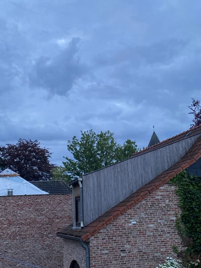 The image depicts a cloudy sky above buildings in Sint-Baafs-Vijve, Belgium. A brick building with ivy and white flowers growing on it is visible, along with a gray wooden roof with red tiles. Another building, partially covered with white plastic sheeting, is also present. A church steeple rises in the background. Deciduous trees with green and maroon leaves fill the scene.