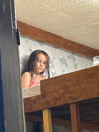 A young girl with dark hair and brown eyes peeks from behind a wooden beam in what appears to be an attic space. She is wearing a pink tank top and her hair is slightly damp, framing her face. The background features patterned wallpaper with blue floral designs. To the left, there is a textured cream-colored wall or ceiling panel. The perspective is looking upwards, with the girl positioned in the center of the frame, slightly to the left. The lighting suggests it is daytime.