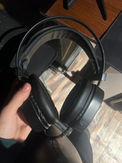 The image depicts a pair of black over-ear headphones being held by a human hand, observed in what appears to be an indoor setting in Tuyabuguz, Uzbekistan.
**Subjects/Objects:**
* **Headphones:** A pair of black over-ear headphones is the primary subject. The headband features a padded, textured strap with visible blue stitching along its edges. The earcups are black and appear to be made of a synthetic leather-like material. A black braided audio cable is visible extending from the left earcup.
* **Hand:** A light-skinned human hand, with clean, unpolished fingernails, holds the left earcup of the headphones. A dark green fabric, possibly a sleeve, is visible at the wrist.
**Actions/Interactions:**
* The hand is actively grasping the left earcup of the headphones, holding them upright.
**Environment:**
* **Background:** The floor visible in the background consists of wooden planks in varying shades of brown and beige. Dark, angular structures, consistent with furniture legs or undersides of a desk/table, are visible above the floor.
* **Lighting:** The scene is moderately lit, with shadows suggesting a directional light source, possibly overhead. The overall impression is an interior space.