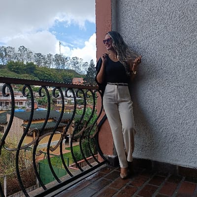 A woman stands on a brick-tiled balcony with an ornate wrought-iron railing, gazing at a cityscape and tree-covered hills under a cloudy sky. She wears a black v-neck top, cream-colored high-waisted trousers, and nude-colored shoes. Her hair is styled in loose waves, and she accessorizes with sunglasses, a gold necklace, and a watch. She leans against a wall, casually touching her hair.