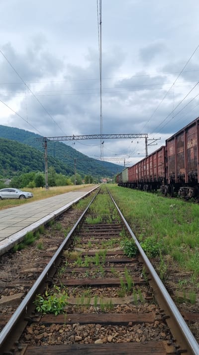 The image depicts a rural train station in Kostryna, Ukraine, under an overcast sky. In the foreground, a set of railroad tracks runs lengthwise towards a distant, hazy horizon, with green grass and weeds growing between the weathered wooden railroad ties and ballast. To the left of the tracks, a concrete train platform is visible, adjacent to a patch of dry grass. A silver sedan is parked on the paved area next to the platform. In the midground, a long line of red and green freight train cars stretches along the tracks on the right side of the image. Beyond the train cars, a densely forested, green mountain range dominates the background, with a few scattered buildings visible at its base. Overhead, power lines and their supporting structures are strung across the sky. The overall scene conveys a sense of quiet solitude and industrial infrastructure integrated with a natural, mountainous landscape.
