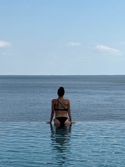 The image displays an individual viewed from the back, seated at the edge of an infinity pool. The subject is wearing a black two-piece swimsuit; the top features an asymmetrical strap design. Their hands rest on the pool's edge, and their reflection is visible in the water. Beyond the pool, a large body of deep blue water extends to a distant, flat horizon line. The sky above is light blue with minimal, faint white cloud formations. The location is Hlibivka, Ukraine.
