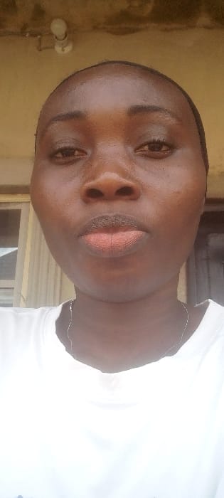 A close-up, front-facing portrait in Agunmo, Nigeria, captures an adult individual with dark skin, visible from the upper chest. The subject wears a white crew-neck t-shirt and a thin, silver-toned necklace. Their dark hair is pulled back, not clearly delineated. Facial features include dark eyes and full lips, which are slightly pursed. The background is out of focus, revealing a light-colored ceiling or overhang structure in the upper left, where an exposed, clear incandescent light bulb is mounted. To the left, a vertical-slatted window or blind is partially visible behind the subject. The ambient lighting appears bright and natural.