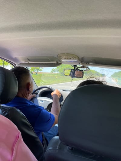 The image is a view from the backseat of a car, looking forward. A man in a blue polo shirt and blue jeans is driving, his hands on the steering wheel. His face is not clearly visible, but a reflection in the rearview mirror shows his face, he is wearing sunglasses. Outside the car, the environment is rural with fields and trees under a clear sky. There are two other passengers in the car, one in a pink top in the foreground and another whose hair is visible in the middle ground. The location is not specified in the image but is identified as Trunovskoye, Russia.