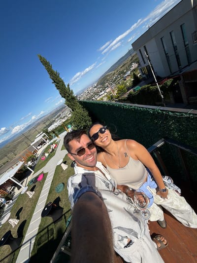 A couple is captured in a selfie taken from a high vantage point overlooking the city of Lisi, Georgia. The man, in the foreground and slightly to the left, wears sunglasses and a patterned shirt. The woman, to his right, also wears sunglasses and a light-colored tank top. She has a tattoo on her shoulder and is holding a glass of white wine. They appear to be on a balcony or terrace.

Below them, a green lawn is dotted with white tables and colorful beanbag chairs, some occupied by other individuals. In the background, a city sprawls across a valley, with buildings and mountains visible. A tall, slender tree stands prominently in the left portion of the frame, reaching towards the clear blue sky. The overall impression is of a relaxed outdoor setting on a sunny day.