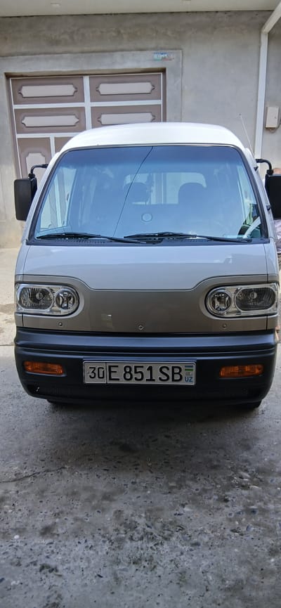 A front-view, medium shot captures a silver minivan parked on a concrete surface in front of a beige garage door. The minivan's headlights, turn signals, and a black bumper are clearly visible. A license plate with the characters "30 E 851 SB UZ" is affixed to the front bumper. The garage door, a neutral beige with darker trim, is partially obscured by the minivan. The overall scene appears to be an outdoor setting in a city. The image is taken at eye level, suggesting the photographer is standing. The lighting suggests it is daytime. The environment is urban, with a building facade and a paved ground.