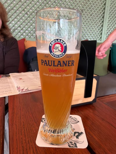 A tall glass of Paulaner Weißbier is centered on a wooden table. The beer is a pale amber color with a frothy head. The glass has the Paulaner logo printed on it, which features a man's profile within a circular emblem. Below the logo, "München" and "1634" are visible, along with the text "PAULANER Weißbier" and "Feinste Münchener Braukunst". The glass rests on a square coaster with a black and white design.

In the background, a lattice-like green wall is visible, suggesting an outdoor or semi-outdoor seating area. To the left of the beer, a portion of a person's dark brown hair and shoulder is visible, along with some colorful cushions on what appears to be a bench. To the right of the beer, a person's arm, wearing a green garment, is partially in view, with their hand reaching towards the right edge of the frame. A black holder containing a wooden object is also present.

A menu is laid out on the table to the left of the beer glass. The overall setting appears to be a restaurant or beer garden. The specific location in the city of Otterlo, Netherlands, is not discernable from the visual elements of the image itself.