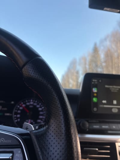The image displays the interior perspective of a moving vehicle, looking forward. In the immediate foreground, the left side of a black leather steering wheel is prominent, featuring red stitching and perforated grip sections. A silver paddle shifter marked with a '+' symbol is visible behind the steering wheel. Through the upper-left spoke of the steering wheel, a portion of the vehicle's instrument cluster is visible, showing a dark background with a red needle indicating a speed between 60 and 70 units. To the right, an infotainment screen displays an active interface, likely Apple CarPlay, featuring Russian text "Москва-Москва" (Moscow-Moscow) and music playback controls. The exterior environment, viewed through the windshield, consists of a clear, light blue sky dominating the upper background, with a blurred line of leafless trees extending across the horizon. This scene is located in the city of Semenovo, Russia.