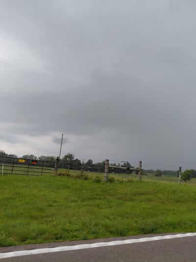 The image depicts a landscape scene with a prominent overcast sky. To the right, a paved road with a white line is visible, suggesting a roadside view. Adjacent to the road is a grassy field that extends into the distance. A fence, constructed with wooden posts and wires, runs along the edge of the field. Further into the field, a black metal gate is present, with two signs attached to it. One sign is orange and rectangular, while the other is yellow and also rectangular. Beyond the field, a line of trees forms the horizon, under the heavy grey clouds. The overall impression is of a rural or semi-rural setting, possibly on the outskirts of Bagdad, United States. The weather appears to be gloomy, with the overcast sky indicating a cloudy or potentially rainy day. The time of day is not explicitly clear but the subdued lighting suggests it could be daytime.