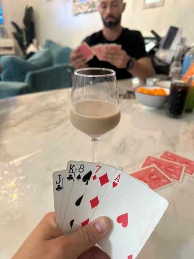 The scene is set indoors within the city of Fort Lauderdale, United States, depicting a leisure activity involving multiple individuals. In the immediate foreground, a human hand holds a fanned arrangement of five playing cards: Jack of Clubs, King of Spades, 8 of Spades, 7 of Diamonds, and Ace of Hearts, with a partial view of a sixth red-suited card. A stemmed glass, approximately half-filled with a beige opaque liquid, stands on a white marble-patterned tabletop. To the right of the cards held by the hand, a stack of four red-backed playing cards lies face down on the table. In the midground, a white bowl containing orange-colored snacks, a glass with a dark liquid beverage, and a green-colored container are visible on the table surface. In the background, a male individual with a beard and a dark short-sleeved shirt is seated, out of focus, holding a fanned hand of red-backed playing cards. Behind the male, light blue upholstered furniture, a dark potted plant, and light-colored walls adorned with decorative elements are present. The individuals are engaged in a card game.