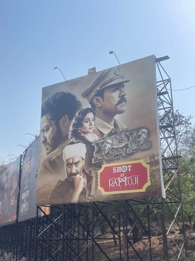 A low-angle, outdoor photograph shows a billboard advertising the film "RRR," shot at Ramoji Film City, located somewhere in India. The billboard is mounted on a metal frame structure. The sky is blue. The billboard features a collage of the movie's actors. Below the "RRR" billboard, another billboard is visible. Trees and foliage can be seen behind the billboard and its support structure.