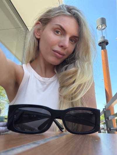 A blonde woman wearing a light pink tank top is taking a selfie outdoors. She has on a pair of black sunglasses which are resting on a wooden table in front of her. Behind her, a tall metal lamp post with a wooden base is visible against a bright blue sky. The woman has a single gold earring in her visible ear. The image appears to be taken during the day in an outdoor setting, possibly a cafe or patio area. The location is identified as Húmera-somosaguas-prado Del Rey, Spain.