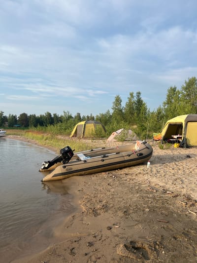 A calm, sandy shoreline in Borovoye, Russia, features a campsite with two tents, one yellow and one pink. An inflatable boat with a black outboard motor is beached on the sand near the water's edge. The surrounding area is covered with trees and vegetation, and the sky is a pale blue with scattered clouds. The scene suggests a peaceful, natural environment, ideal for outdoor recreation.