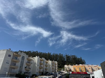 The image displays a bright, sunny daytime scene in a residential area, likely Montemor-o-Velho, Portugal. In the foreground and midground, several multi-story apartment buildings with predominantly white and light yellow facades and balconies line what appears to be a sloping road or hillside. Numerous cars are parked along the street and in parking areas.
Behind these buildings, a dense, verdant forest of tall, slender trees covers a steep hill, creating a natural backdrop against the sky. The sky is a vibrant blue, sparsely dotted with delicate, wispy white cirrus clouds, indicating fair weather. A small, dark bird is visible in flight in the upper left section of the sky.
To the right, a series of more colorful, two-story buildings, painted in shades of yellow and orange, are situated above a commercial area. Visible signage includes "Jet-Wash," "Aspiração" (likely referring to vacuum services), and "Intermarché," indicating a car wash and a supermarket or related retail establishment. A person in work attire is partially visible in the lower left foreground near a parked car. The strong sunlight creates clear shadows, emphasizing the architecture and the varied terrain. The scene depicts a typical, peaceful day in a residential and commercial locale.