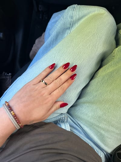 A close-up, high-angle shot captures a person's hand resting on their lap, adorned with vibrant details. The hand, with fair skin, features long, almond-shaped fingernails painted with a glossy, deep red polish. On the ring finger, a gold-colored band ring with a repeating heart-shaped pattern is visible. The wrist is accessorized with two bracelets: an upper bracelet made of small, round beads in shades of brown, orange, and yellow, and a thinner, lighter-colored (possibly beige or grey) woven or cord bracelet beneath it. A small, subtle tattoo resembling a simple smiley face is visible on the back of the hand, closer to the wrist.
The person is wearing light-wash denim jeans, which appear to be folded or creased, creating texture on the fabric. A darker, possibly brown, garment or bag is partially visible on the right side of the frame, contrasting with the lighter denim. The lighting is bright and even, highlighting the details of the hand and clothing. The overall scene suggests an indoor or car setting, with dark, indistinct shapes in the bottom left corner, possibly indicating a car interior or furniture. There is no visible text in the image. The image provides a detailed view of the hand and personal accessories without revealing the person's face or specific activity, focusing instead on fashion and stylistic choices.