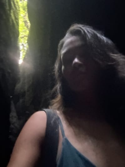 The image shows a close-up, low-angle shot of a person's head and shoulder. The person has long, dark hair that is blurred due to motion. The lighting is dim, with a bright, out-of-focus green and yellow light source visible in the lower left portion of the frame, suggesting a natural setting possibly illuminated by a flashlight or similar device. The background is mostly dark and indistinct. The person appears to be wearing a dark-colored garment, possibly a t-shirt, on their shoulder. The overall impression is intimate and slightly mysterious, with the focus on the texture of the hair and the play of light. There is no visible text in the image. Given the location context of Bangli, Indonesia, the scene could be interpreted as a person in a natural environment at night, perhaps exploring or camping. The blurred motion suggests movement, possibly a selfie taken while active or in low light conditions.