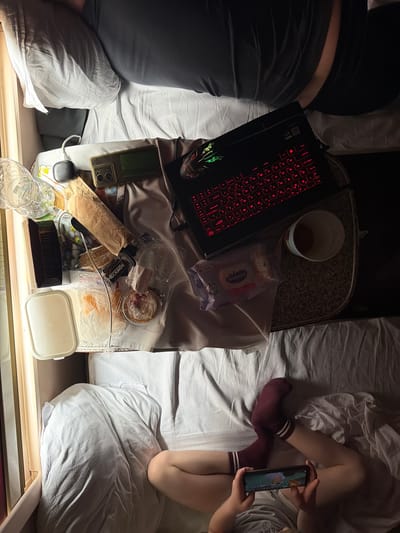 An overhead, low-light shot captures a cozy, confined sleeping area, likely a train compartment or small room, in Bobrov, Russia. Two individuals are resting on separate beds with white bedding, separated by a narrow table or shelf laden with various items.

On the left, a person is seen from the back, lying on their side, wearing dark clothing. Their head and upper body are out of frame. Adjacent to them, on the table, is an open dark laptop with a red-backlit Cyrillic keyboard, next to which sits a small potted plant and a gold-colored smartphone with a green case, accompanied by a computer mouse.

On the right, another person is reclined, wearing maroon socks with white stripes. They are actively engaged with a smartphone, holding it in both hands and looking at the screen, which displays what appears to be a game or video.

The table between them serves as a cluttered eating and working space. Items include a cup with a dark beverage, a package of "Watashi" brand wet wipes featuring a cartoon rabbit, and an assortment of snacks. These snacks are in clear plastic containers and bags, including what looks like pastries or cakes (one with a red topping) and a paper-wrapped item, possibly bread. Visible text on packaging includes "НЕКСКАФЕ" (NESCAFE in Cyrillic, possibly a brand for a food item) and "СОСТАВ" (ingredients). A clear plastic water bottle and charging cables are also present.

The scene suggests an evening or nighttime setting, with both individuals engaged in personal entertainment and consuming snacks in a relaxed, intimate atmosphere.