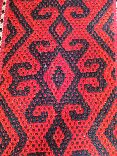 The image is a close-up, eye-level shot of a textile with a geometric pattern in red and black. The pattern consists of repeating diamond shapes and swirling motifs formed by the arrangement of red and black stitches. The texture of the textile is visible, with individual threads creating a slightly raised, pixelated effect. The lighting appears to be natural, casting soft shadows that highlight the texture. The image is a detail of a larger item, possibly a rug, tapestry, or garment. No specific location in Batken, Kyrgyzstan is identifiable from the image itself.