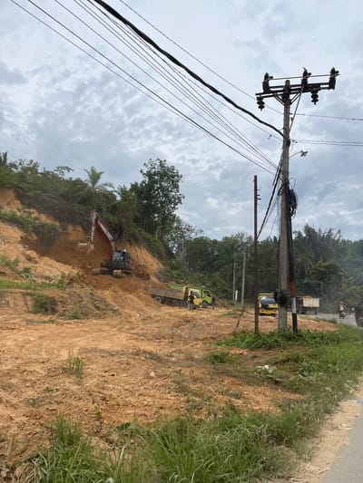 This media file depicts a construction or excavation scene in Rantau Pauh, Indonesia. The primary subjects are heavy machinery, including an excavator and two trucks, actively engaged in moving soil. The excavator is positioned on a dirt embankment, its arm extended and bucket full of earth. One truck is parked on a higher level of the embankment, likely ready to receive material, while another truck is visible in the background on a road. Several utility poles with numerous wires are prominent in the foreground, suggesting the work is being done near a developed area or infrastructure. The scene is set on a steep, raw, earth embankment with patches of green vegetation growing on it and on the surrounding hillsides. The sky is overcast with visible clouds, indicating a cloudy day, possibly with a chance of rain. The time of day appears to be daytime, given the lighting conditions. The overall impression is one of ongoing earthwork and infrastructure maintenance or development. No visible text is discernible in the image.