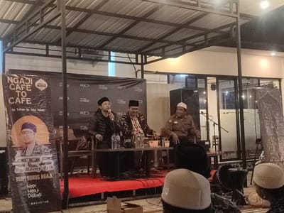 The scene takes place at the "Ngaji Cafe To Cafe" in Kecamatan Pandaan, Indonesia, located at coordinates -7.653 latitude and 112.688 longitude. Three men are seated on a stage, facing the audience. The man on the left is speaking into a microphone. He is wearing a black jacket, black pants, and a black cap. The man in the middle is wearing a brown jacket, brown pants, and a black cap. He is gesturing with his right hand. The man on the right is wearing a brown shirt, brown pants, and a white cap. He is smiling and looking towards the left. All three men are sitting behind a table, with two glasses and a water bottle on the table. The stage is covered with a red carpet and a black backdrop with white writing. The backdrop features the cafe's logo and name. There is a large, white sign to the left of the stage that reads "Ngaji Cafe To Cafe." The sign features an image of a man in a brown shirt and white cap, with a yellow sun behind him. The writing on the sign states "Se Tuhan Se Jala Jalan." The sign also features the phrase "Gus Romy Style" and "NGOPI BONUS NGAJI." Another sign on the wall features the phrase "Noji Diskus." There is a table and chairs to the right of the stage. An audience member in a white cap is seated behind a black speaker in the front row. The scene is lit by artificial lighting, creating a warm glow. The scene is indoors, with an open roof and a partial view of a building beyond. The building has large windows, and the windows are covered with glass.