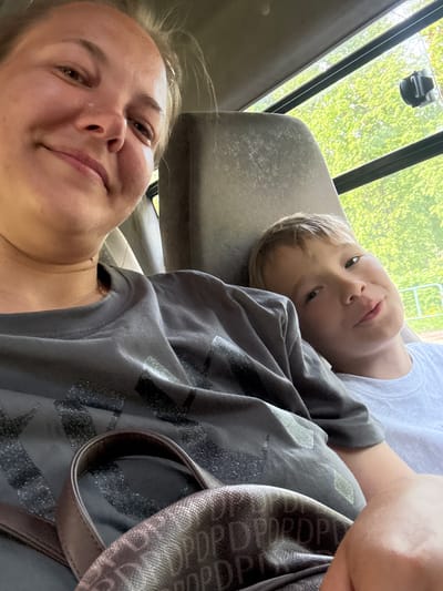 A woman and a young boy are inside a vehicle, possibly a van or bus, in Novosinkovo, Russia. The woman is in the foreground, partially out of focus, looking toward the camera. The boy is lying on the back of the seat. Both are wearing casual clothing. A patterned bag is partially visible. Outside the vehicle, there are trees.