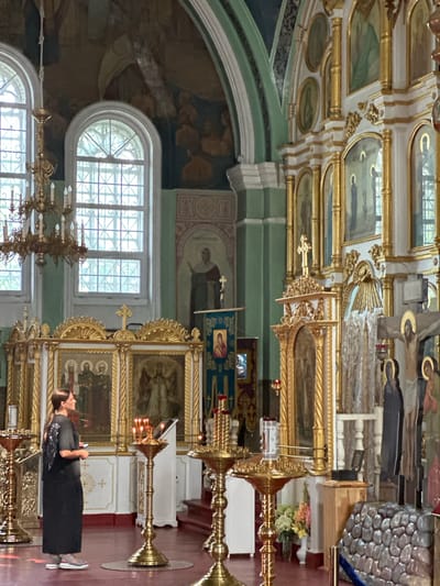 The image depicts the interior of a church, possibly located in Nogornyi, Russia. A woman in a black dress and gray shoes stands to the left of the frame, holding a phone in her left hand. She is facing a tall, ornate golden iconostasis, which features multiple religious icons and golden columns. In front of the iconostasis are several golden candle stands with lit candles. The church's interior is richly decorated with religious artwork on the walls and ceiling, and large arched windows with intricate metal grilles are visible. The floor is a deep red color. The overall atmosphere suggests a religious service or a visit to a place of worship.