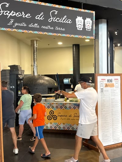 In a bustling Italian pizzeria, likely in Carini, Italy, a family is captured in a playful moment, attempting to mimic a plank or a similar exercise pose. The scene unfolds in front of a traditional wood-fired pizza oven, adorned with a mosaic pattern, hinting at the authentic Italian culinary experience offered. Above, a menu board, framed in wood, displays various Italian dishes, including "Antipasti," "Primi," "Secondi," "Insalatone," and "Pizze." A prominent sign on the left, featuring the logo "Sapore di Sicilia" with stylized portraits, further emphasizes the Sicilian theme. The lighting suggests an indoor setting, possibly during daylight hours. The people appear relaxed and engaged in a lighthearted activity, creating a lively and family-friendly atmosphere. The pizzeria's interior features dark tiled walls and stainless-steel elements, adding to the functional yet inviting ambiance.