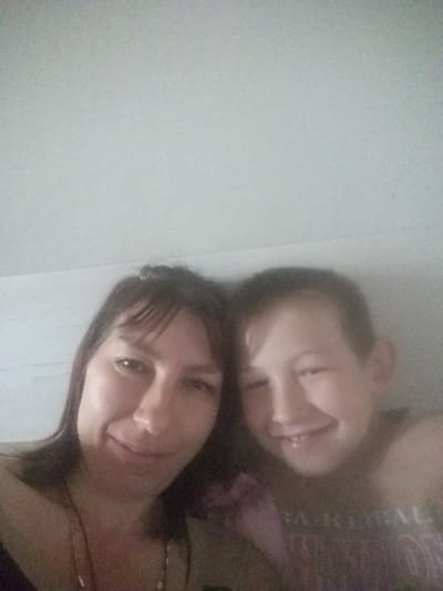 A selfie shows a woman and a child, possibly a mother and son, indoors. The woman is at the top of the frame with brown hair and bangs, smiling slightly. The child is below her, also smiling. The image appears slightly blurry and grainy. The background is a plain, light-colored wall. There is no specific information to determine the location is within the city of Бородулиха ауылдық округі, Kazakhstan, based on the visual content alone.