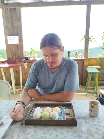 In Trường An, Vietnam, a person sits at a table covered with a floral tablecloth. In front of them is a wooden tray holding several scoops of ice cream in various colors, including white, pale yellow, and pale green. A small silver spoon rests in the tray, and the person's left hand, adorned with bracelets, is reaching towards it. To the right of the tray is a ceramic cup with a brown, abstract pattern. The background shows a wooden structure with large windows offering a view of a lush, green landscape with rolling hills under a bright sky. A sign on the wooden wall reads "TIỆM KEM TRONG RỪNG" in white text on a dark background. Two small round tables, one red and one with a white base, are visible on the left, and a multi-colored stool stands on the right. The overall scene suggests an outdoor cafe or restaurant.