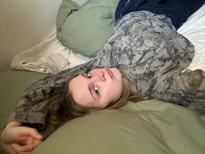 A person lies on a bed with green sheets and a matching pillow, looking directly at the camera. The person's head rests on the pillow, and the body stretches across the bed at an angle. A patterned gray and black long-sleeved shirt is visible. Dark blue pants are also visible. A white pillow and a fluffy blanket are behind the person, adding to the relaxed, casual setting. The overall composition suggests a candid, informal moment.