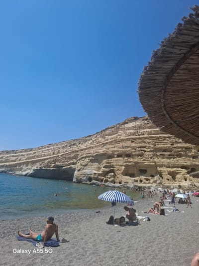 A panoramic, eye-level view of a pebble beach under a clear blue sky, with a large thatched umbrella in the foreground and a steep, layered cliff face overlooking the sea in the background. The beach is populated with sunbathers and swimmers. A person wearing turquoise swim trunks and a cap sits on a towel in the left foreground, facing away from the camera, with shoes placed beside them. Further back, several individuals are visible on the beach and in the shallow, clear blue water. The cliff face, a dominant feature, exhibits numerous caves and crevices. The scene appears to be located at Matala Beach in the Tybakio Municipal Unit, Greece, known for its historical caves and vibrant beach scene.