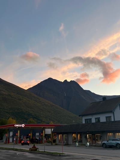 This image depicts a twilight scene in Nordkjosbotn - Gárgán, Norway. In the foreground on the left, a Circle K gas station features a red canopy with the illuminated "CIRCLE K" logo. Multiple fuel pumps are visible beneath the canopy, with a dark-colored sedan, its taillights illuminated, positioned at one pump. Another dark vehicle is partially visible further along the pump line, and one individual is discernible near the pumps. Adjacent to the gas station on the right is a single-story commercial building with a dark, tiled roof, a light-colored facade, and several internally illuminated windows. Above this, a two-story white building with a dark roof and multiple windows is partially visible. A light blue SUV is parked on the road in front of the commercial building. The background is dominated by two mountains: the left mountain is heavily forested with dark green foliage, while the right mountain is rocky, dark gray, and exhibits visible light-colored streaks. The sky above displays a gradient from light blue at the zenith to pink and orange hues near the horizon, with scattered clouds reflecting these colors.